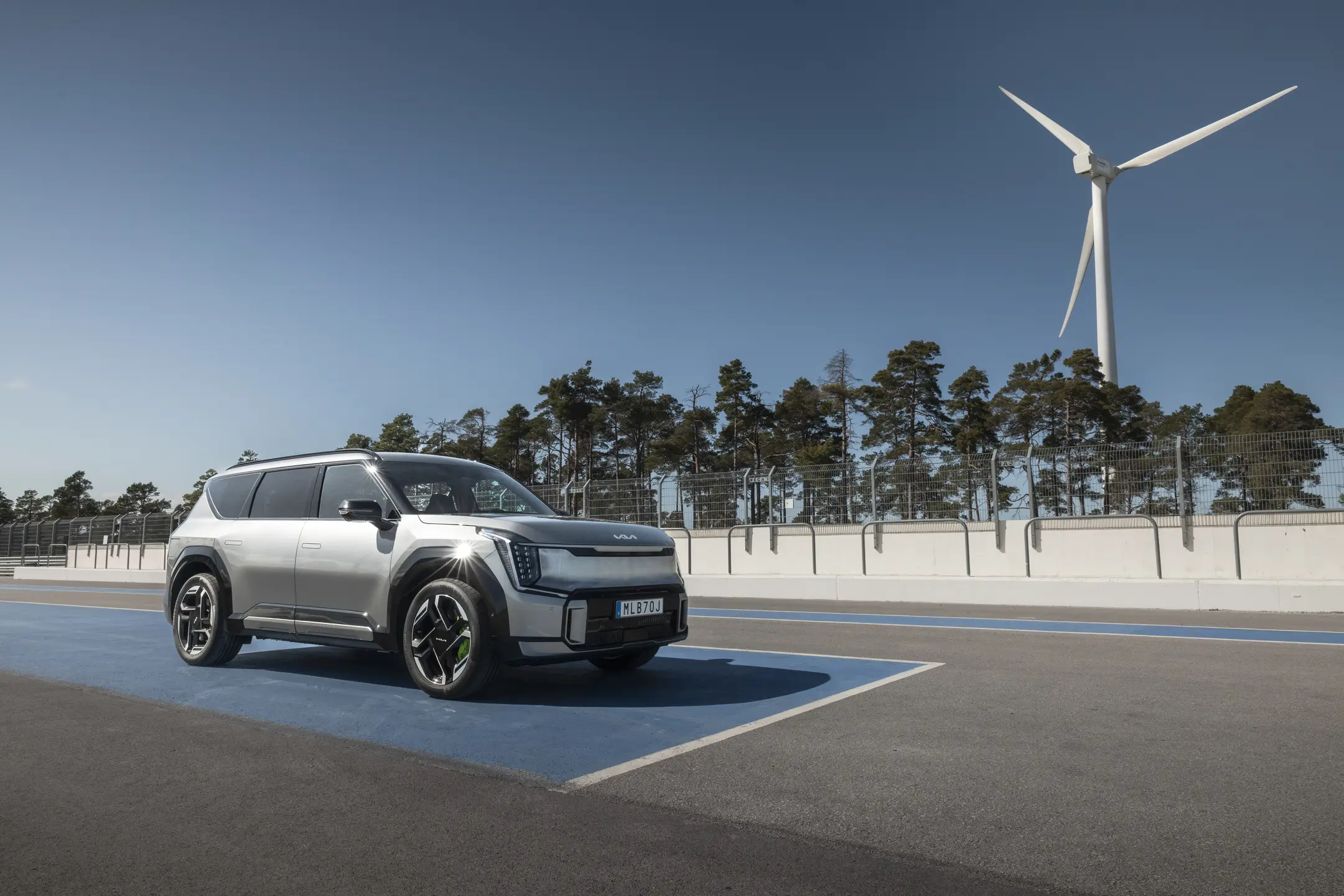 Grå Kia EV6 GT elbil parkerad på blå parkeringsplats framför stora vita vindkraftverk mot klar blå himmel.