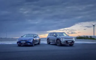 Kia EV6 GT och EV9 GT parkerade sida vid sida vid havet i skymningen, med moln på himlen och vindkraftverk i bakgrunden.