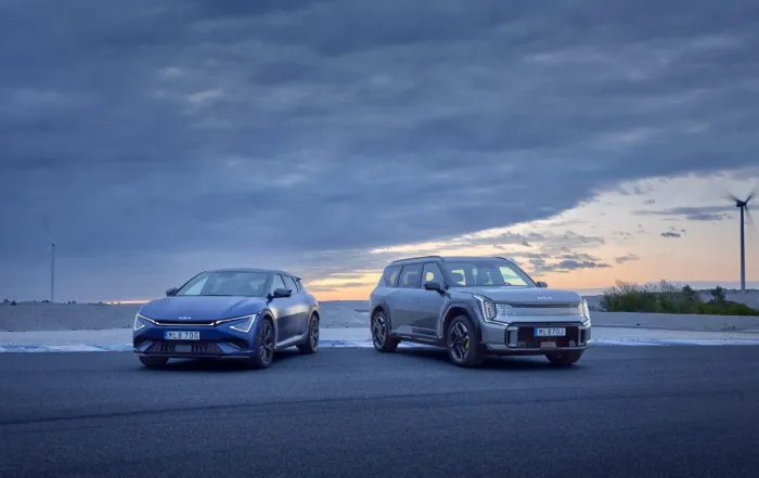 Kia EV6 GT och EV9 GT parkerade sida vid sida vid havet i skymningen, med moln på himlen och vindkraftverk i bakgrunden.