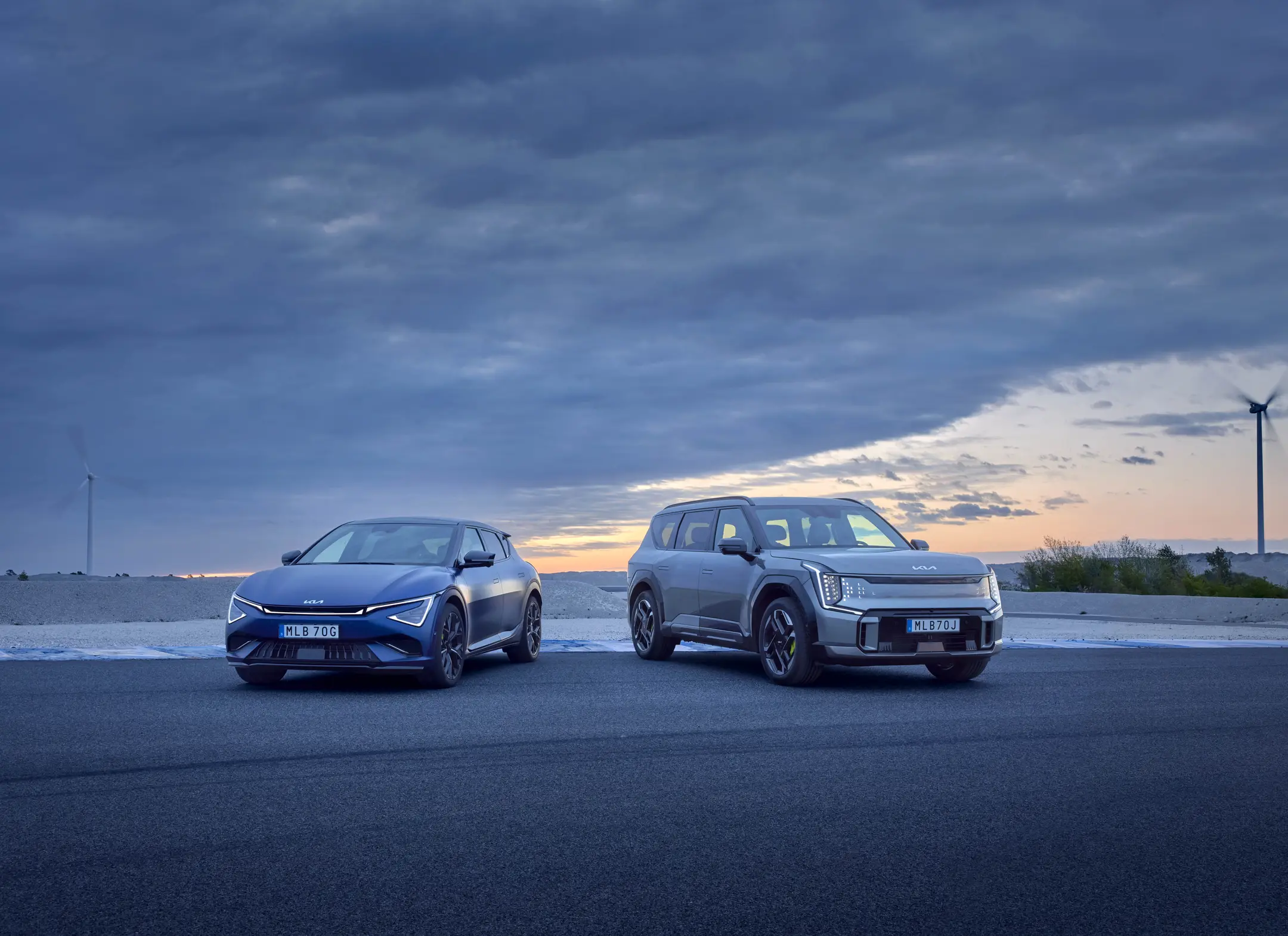 Kia EV6 GT och EV9 GT parkerade sida vid sida vid havet i skymningen, med moln på himlen och vindkraftverk i bakgrunden.