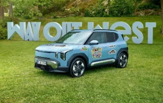 Ljusblå Kia EV2 med Way Out West-stickers parkerad på gräs framför stora bokstäver som bildar Way Out West, omgiven av grön natur