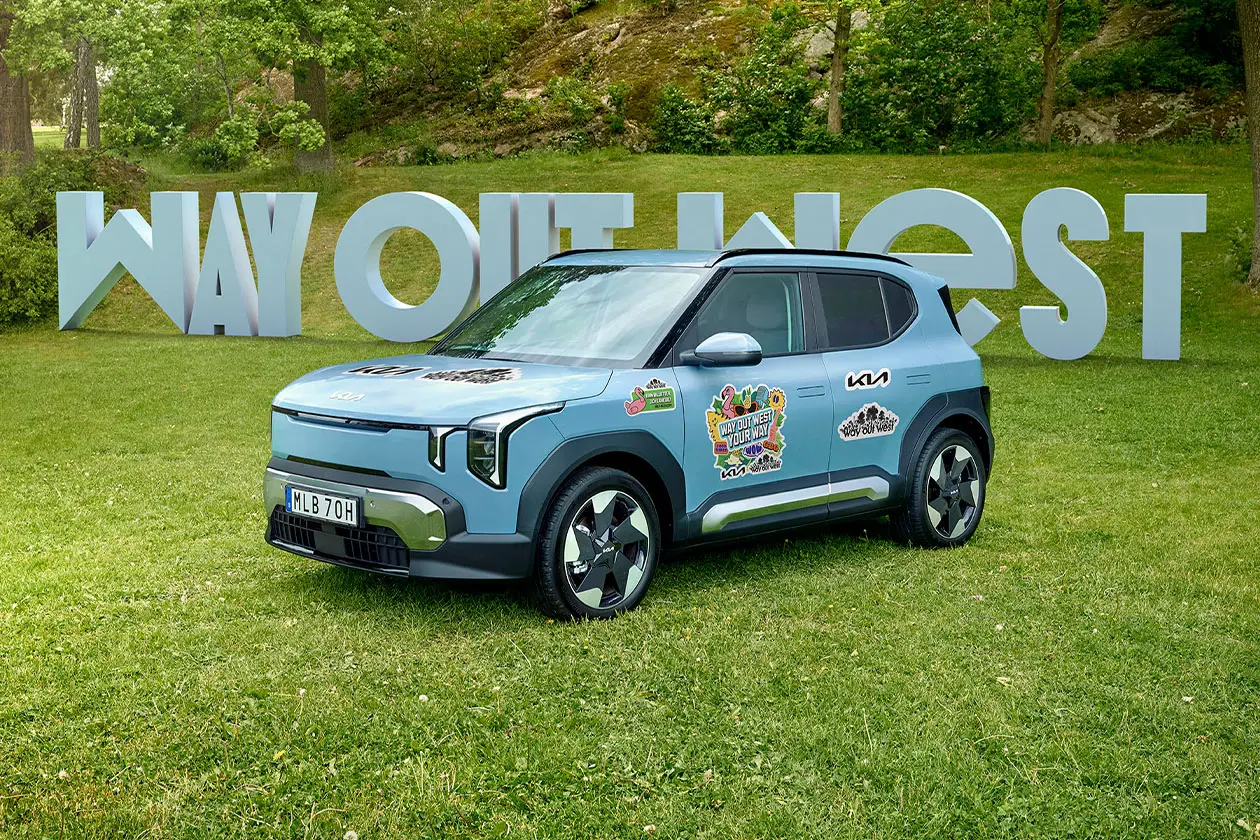 Ljusblå Kia EV2 med Way Out West-stickers parkerad på gräs framför stora bokstäver som bildar Way Out West, omgiven av grön natur